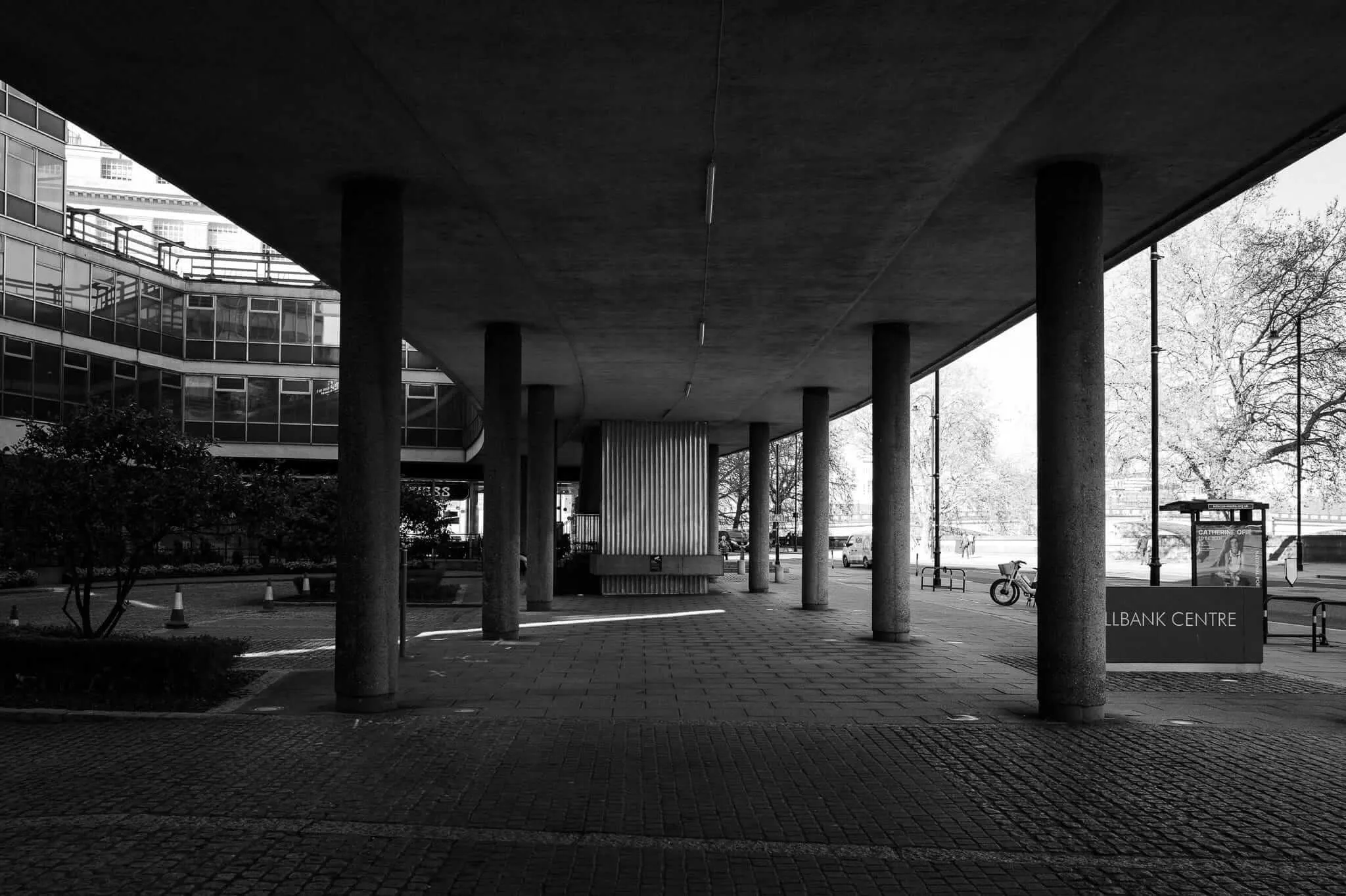 Black and white photograph showing a covered walkway with concrete columns supporting a flat ceiling. The space features a geometric paved floor and opens to views of modernist buildings and bare trees. A sign reading 'LLBANK CENTRE' is visible on the right, and bicycles can be seen parked in the distance.