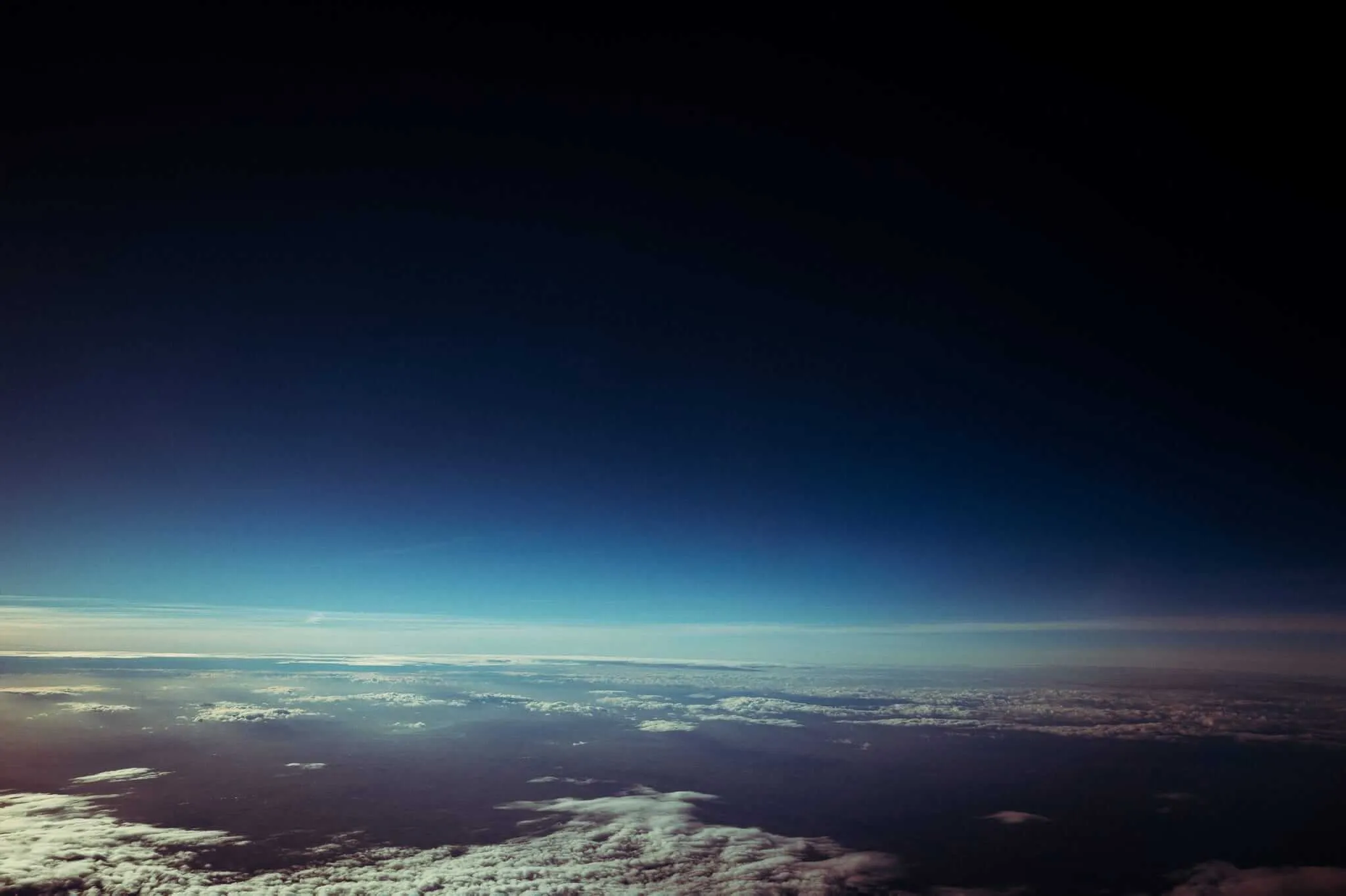 Aerial view of Earth's curvature from high altitude showing the thin blue atmosphere layer transitioning from bright blue sky to deep space black, with scattered white clouds and landmasses visible below