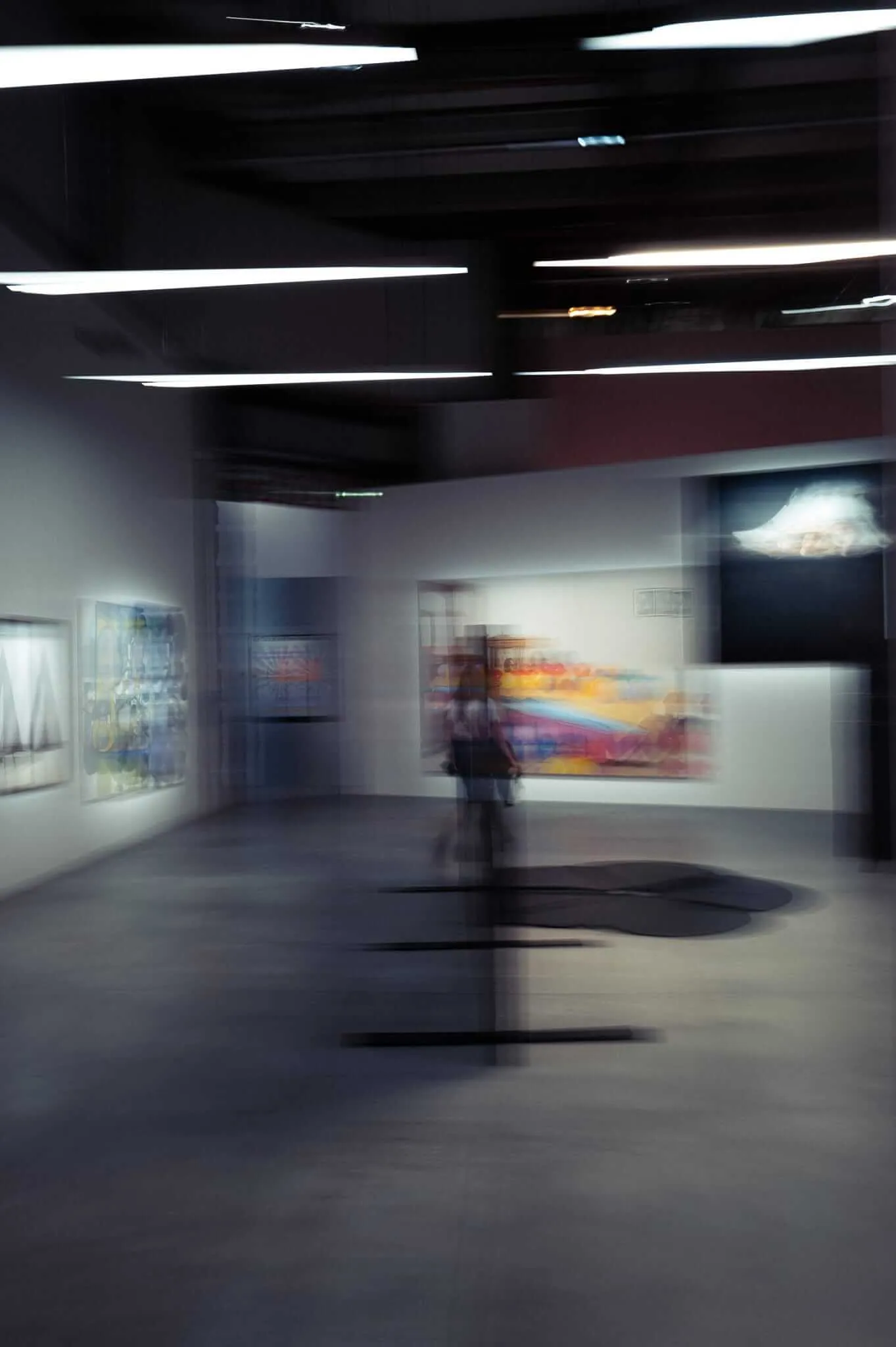 A motion-blurred photograph of an art gallery interior showing a ghostly figure moving through the space, with colorful paintings on white walls, fluorescent ceiling lights, and a dark circular element on the floor