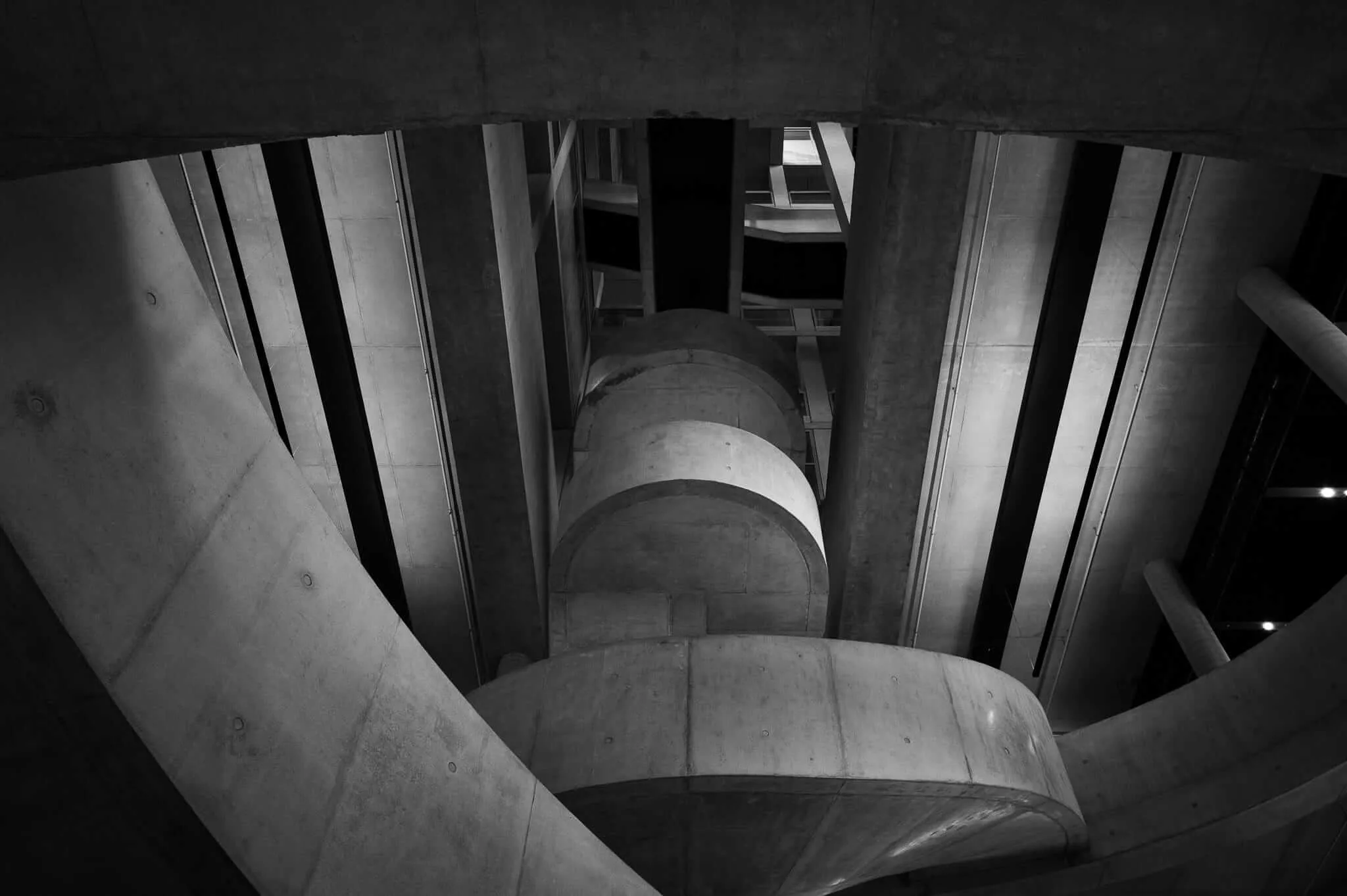 Black and white photograph taken from below showing a dramatic upward view of curved concrete steps or levels ascending in a spiral pattern, framed by vertical concrete pillars and glass panels, creating strong geometric shadows and architectural depth