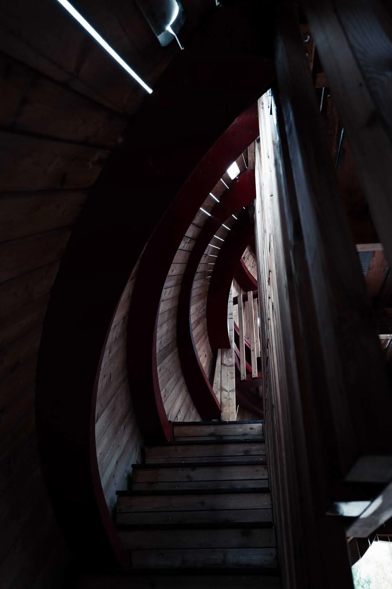 A dramatic low-light view looking up through a wooden spiral staircase with dark red curved railings, receding into a dim tunnel-like structure with minimal lighting visible at the top