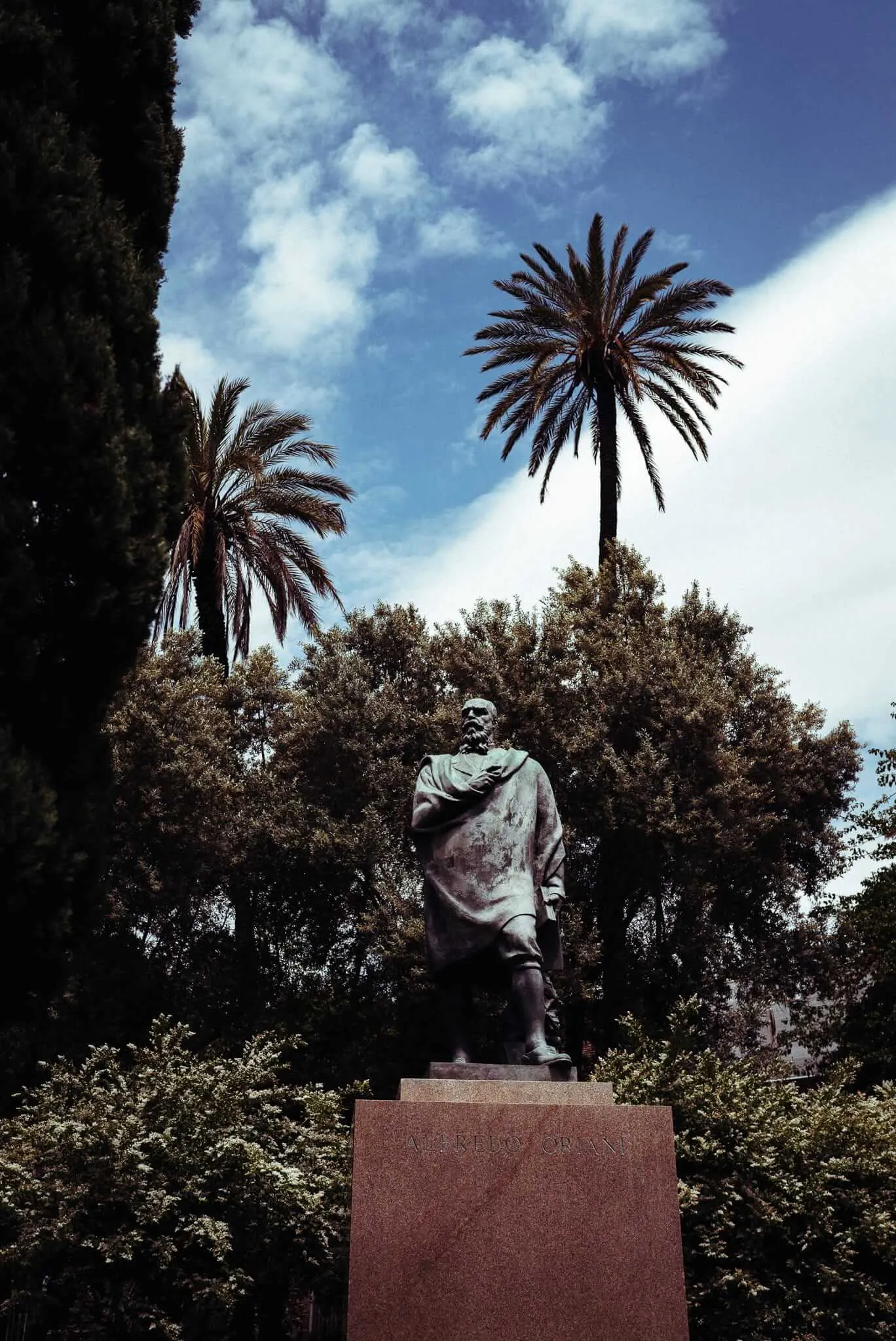 A bronze statue of a robed figure stands on a pedestal against a backdrop of palm trees and blue sky with white clouds, photographed from a low angle that emphasizes the monument's stature