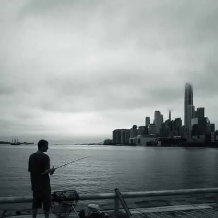 A man stands fishing from a waterfront pier in black and white, holding a fishing rod over calm water, with a dramatic cloudy sky above and a dense city skyline including a prominent skyscraper visible across the harbour. A wire fishing trap and gear rest nearby on the dock.