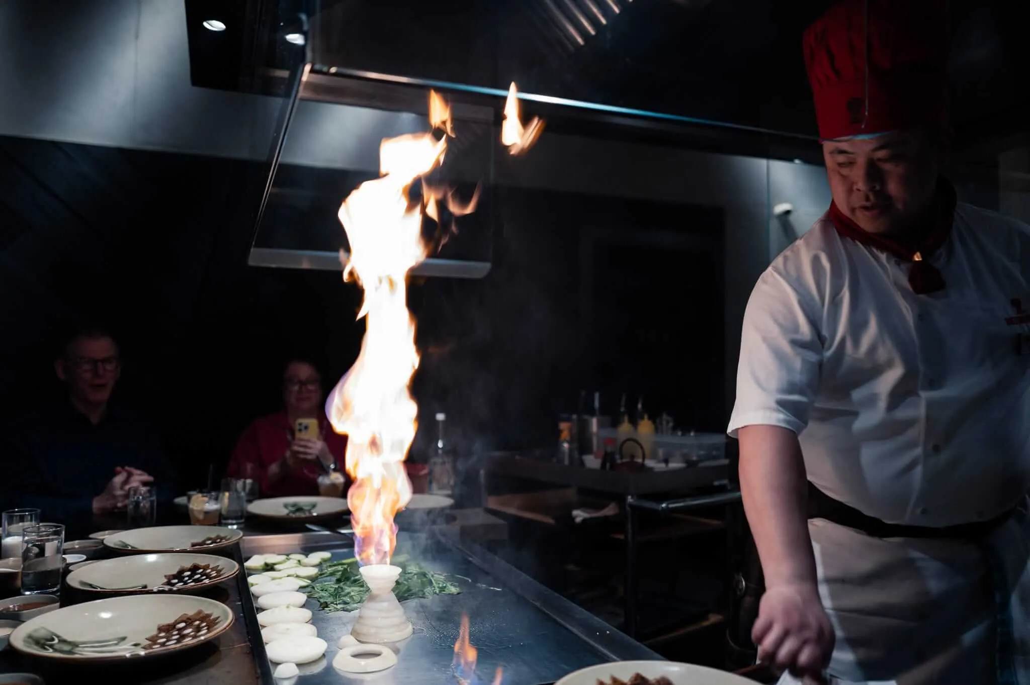 A chef in white uniform and red hat performs teppanyaki cooking at a restaurant, creating dramatic flames on the griddle while customers watch from surrounding tables in dimly lit dining room