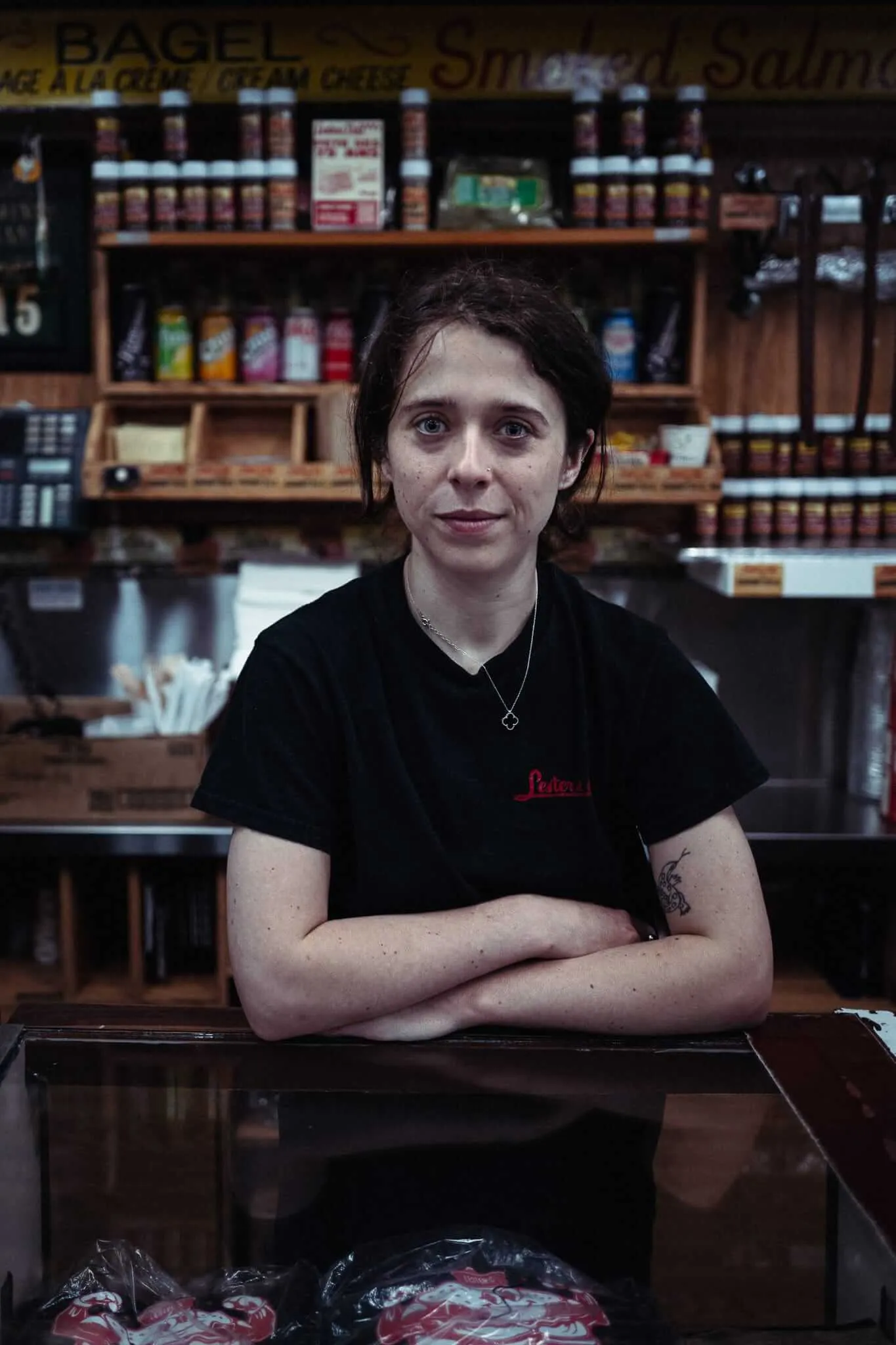 A young woman with dark hair pulled back, wearing a black staff t-shirt and a delicate necklace, leans with arms crossed on a glass deli counter inside a bagel and smoked salmon shop, shelves of jars and bottles visible behind her, looking directly at the camera with a calm, knowing expression.