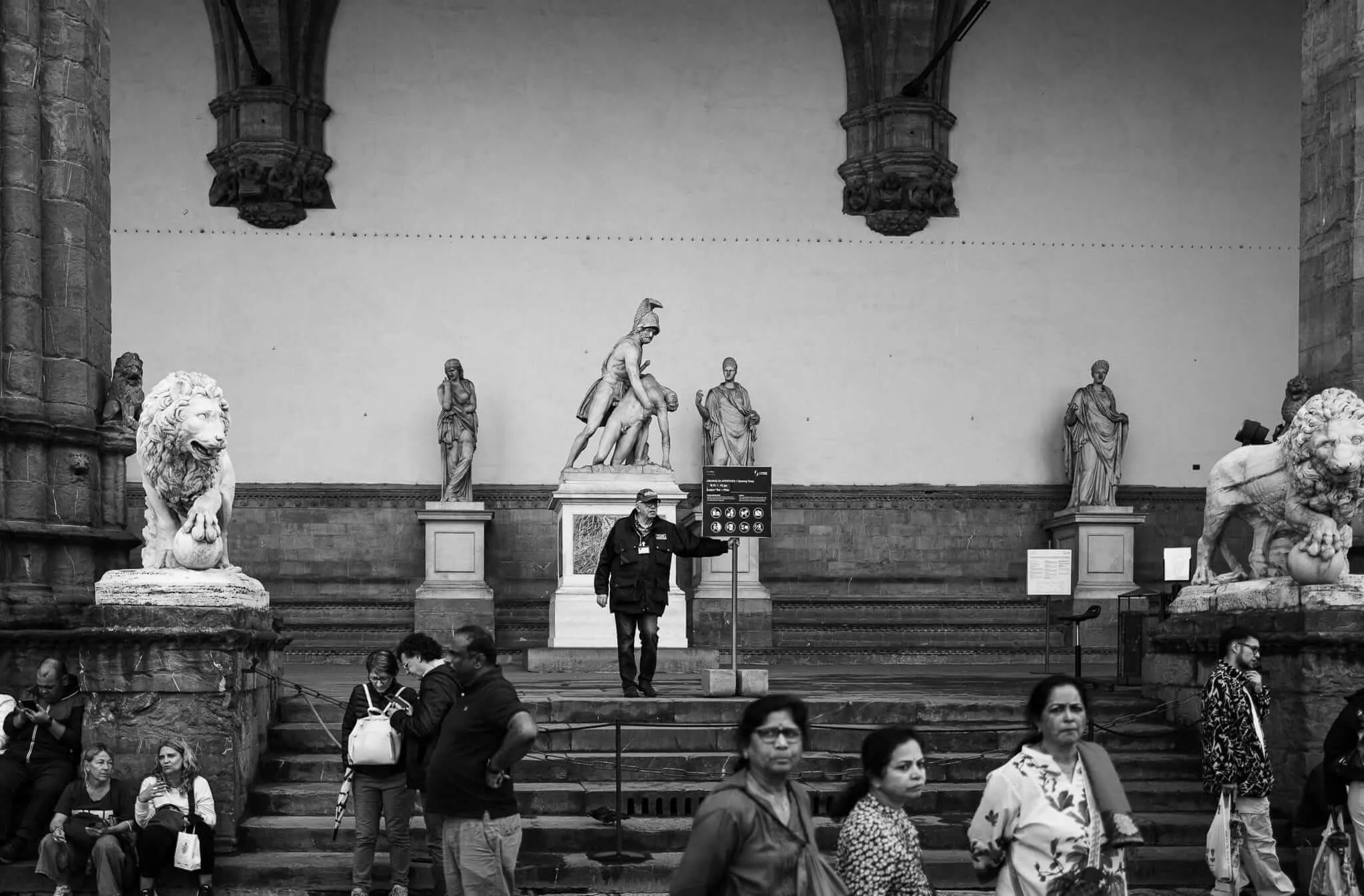 Black and white photograph of tourists gathered at the Loggia dei Lanzi in Florence, Italy, where a security guard stands on the steps pointing toward an information sign, surrounded by Renaissance sculptures including lions and classical figures, while visitors of diverse backgrounds mill about in the foreground.