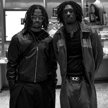 Black and white photograph of two young men with dreadlocks standing side by side in a museum gift shop. The man on the left wears glasses and a leather jacket, while the man on the right wears a longer coat with text on it. Behind them are illuminated storefronts and display cases. The composition creates a sense of solidarity and shared presence in an urban commercial environment.
