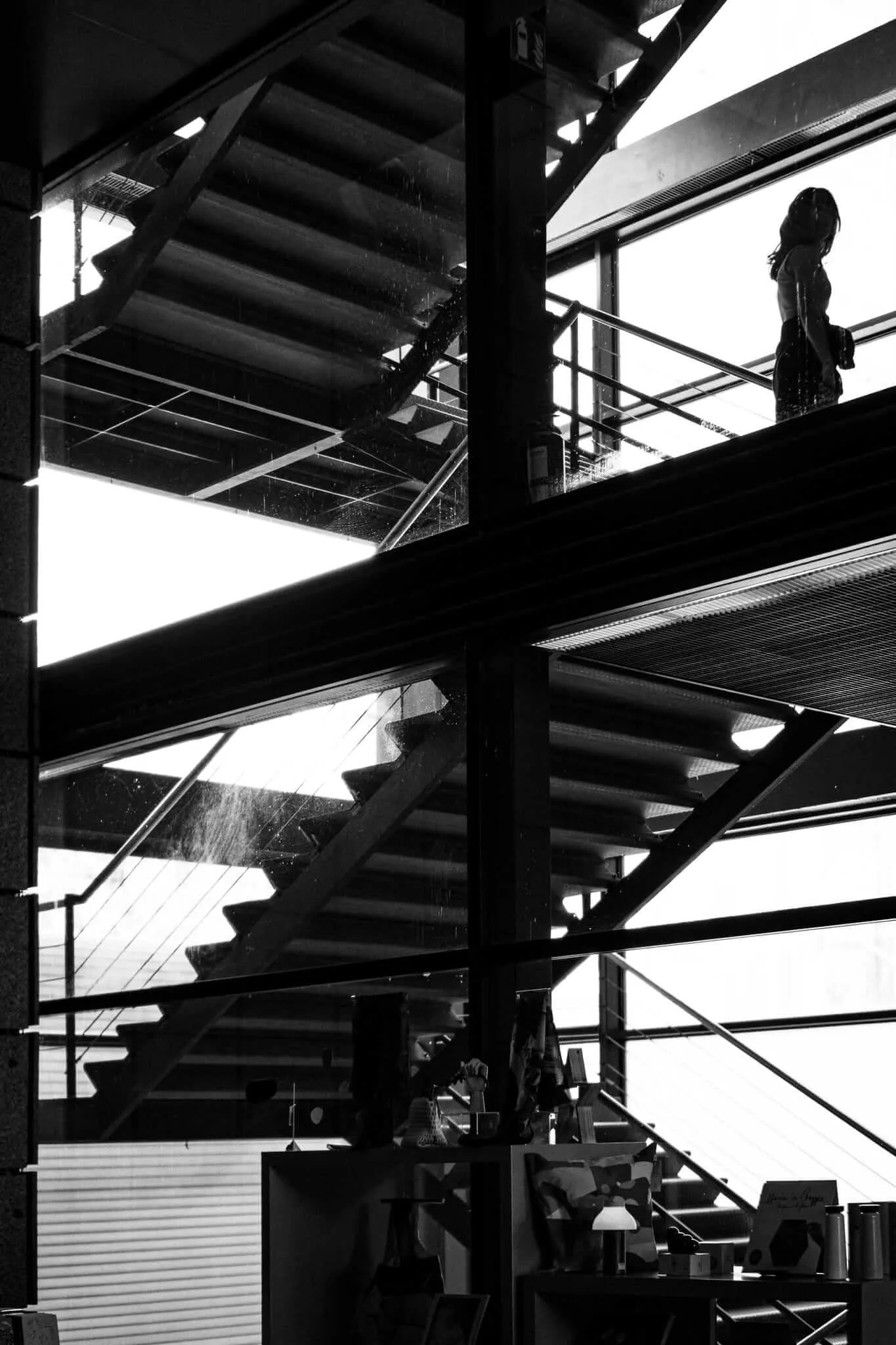 A black and white photograph looking up through multiple levels of an industrial staircase structure. A lone silhouetted figure stands on an upper mezzanine level, backlit by bright light filtering through glass panels. Diagonal steel beams, metal railings, and zigzagging stair undersides create a complex geometric composition.