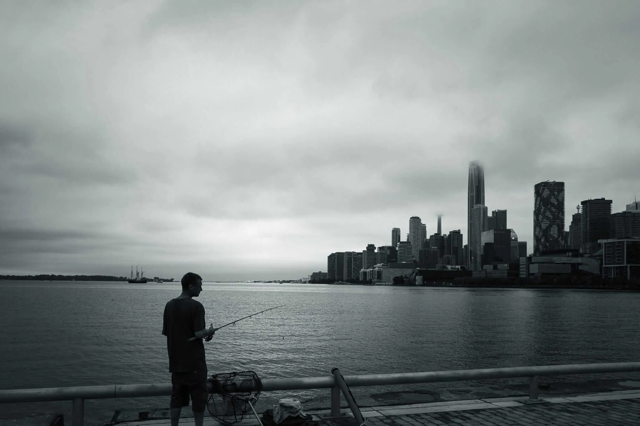 A man stands fishing from a waterfront pier in black and white, holding a fishing rod over calm water, with a dramatic cloudy sky above and a dense city skyline including a prominent skyscraper visible across the harbour. A wire fishing trap and gear rest nearby on the dock.