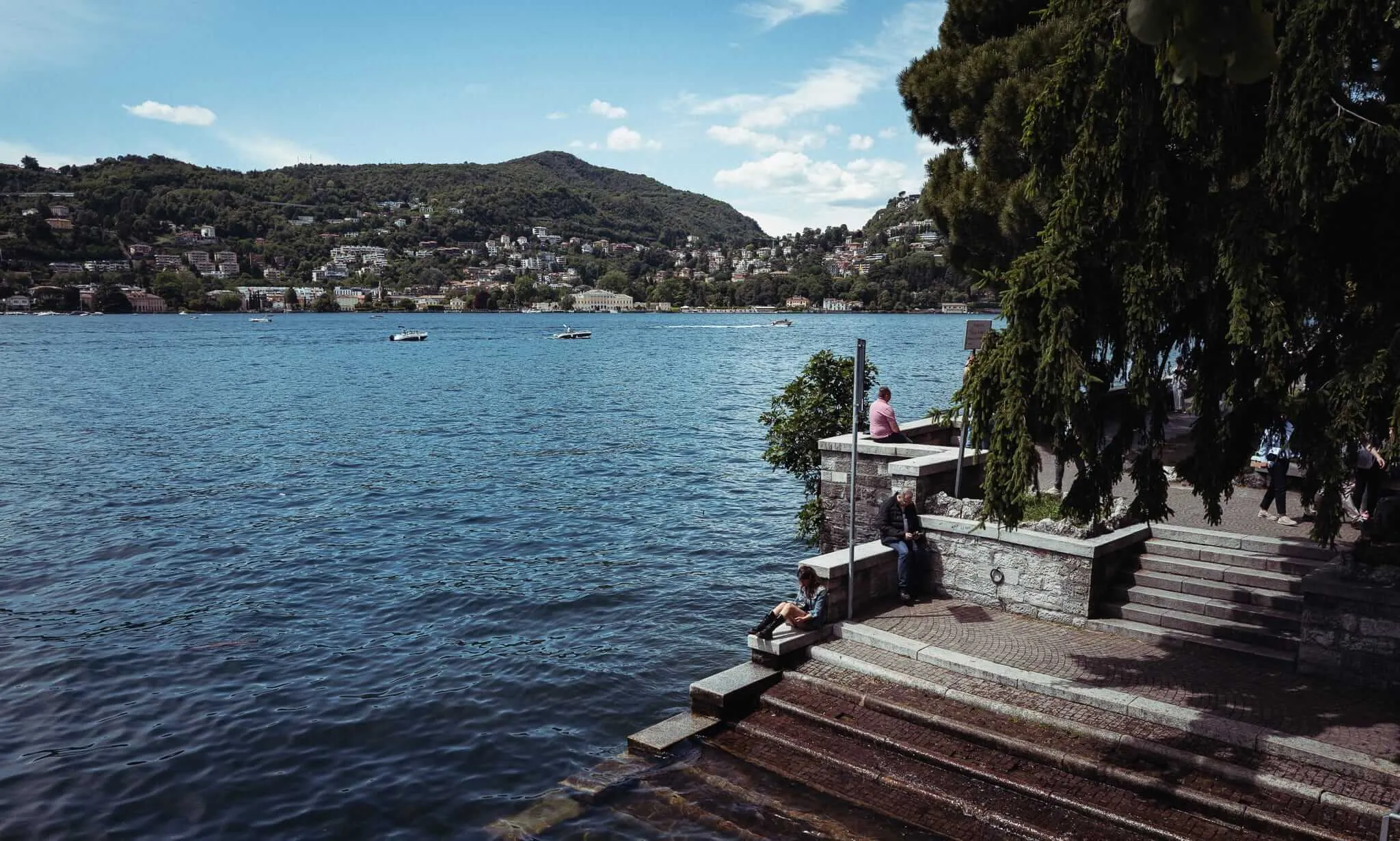 People resting on stone steps and walls along a lakeside promenade, with boats on the water and a hillside town visible across the lake under a partly cloudy sky