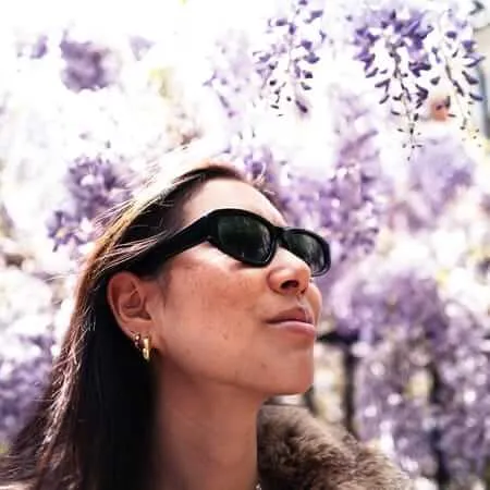 A woman wearing dark sunglasses and gold jewelry looks upward with her face tilted toward the sky, surrounded by blooming purple flowers that create a dreamy, overexposed background